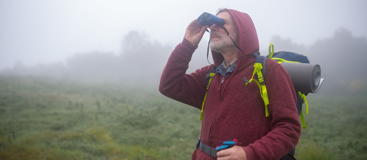 Senior man hiking in foggy landscape, wearing a hoodie and backpack, using binoculars.