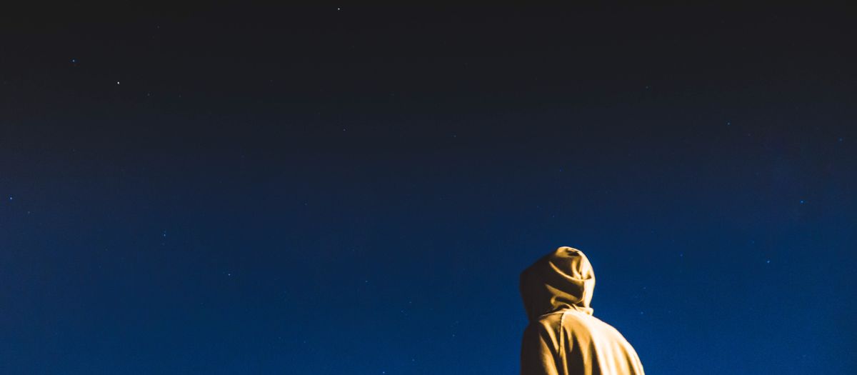 A person on a hillside scanning a star-filled night sky with binoculars on a tripod.