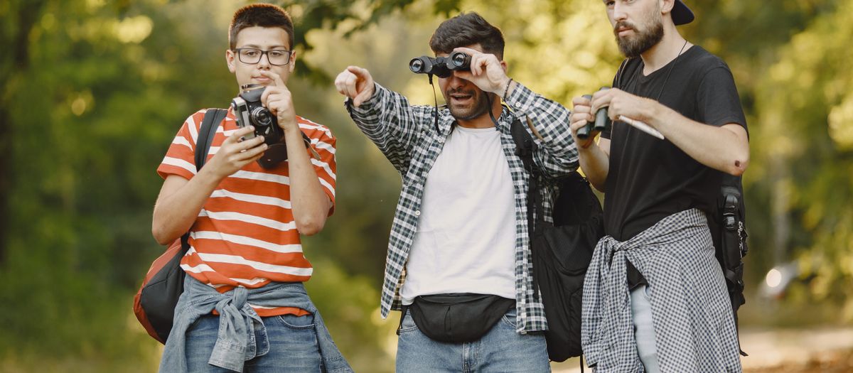 Three men enjoying a hiking adventure in a forest, using binoculars to explore nature.